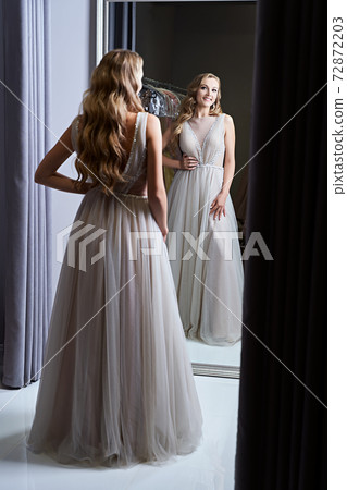 Young beautiful blonde girl wearing a full-length silver white chiffon prom ball gown decorated with sparkles and sequins. Model in front of mirror in a fitting room at dress hire service. Young beautiful blonde girl wearing a full-length silver white chiffon prom ball gown decorated with sparkles and sequins. Model in front of mirror in a fitting room at dress hire service. 72872203