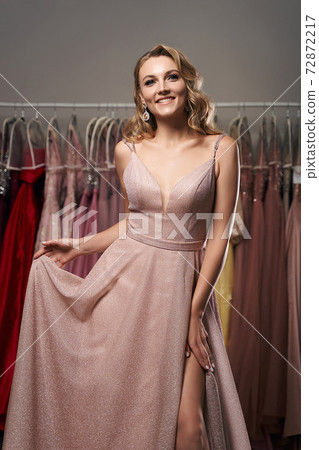 Young beautiful blonde girl wearing a full-length pale pink glitter chiffon draped prom ball gown. Model selecting an outfit for occasion in dress hire service with many options on background. Young beautiful blonde girl wearing a full-length pale pink glitter chiffon draped prom ball gown. Model selecting an outfit for occasion in dress hire service with many options on background. 72872217