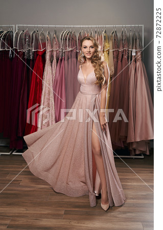 Young beautiful blonde girl wearing a full-length pale pink glitter chiffon draped prom ball gown. Model selecting an outfit for occasion in dress hire service with many options on background. Young beautiful blonde girl wearing a full-length pale pink glitter chiffon draped prom ball gown. Model selecting an outfit for occasion in dress hire service with many options on background. 72872225