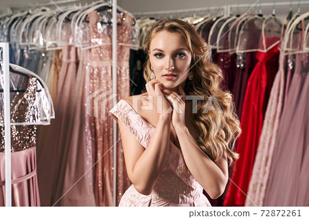 Young beautiful blonde girl wearing a full-length draped one shoulder pale pink satin slit prom ball gown decorated with embroindered pattern. Dress rental service with many dresses in on background. Young beautiful blonde girl wearing a full-length draped one shoulder pale pink satin slit prom ball gown decorated with embroindered pattern. Dress rental service with many dresses in on background. 72872261