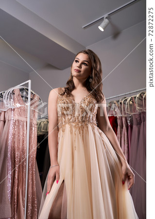 Young beautiful brunette girl wearing a full-length beige yellow champaign chiffon slit prom ball gown decorated with golden sparkles and sequins. Dress hire service with many dresses on background. Young beautiful brunette girl wearing a full-length beige yellow champaign chiffon slit prom ball gown decorated with golden sparkles and sequins. Dress hire service with many dresses on background. 72872275
