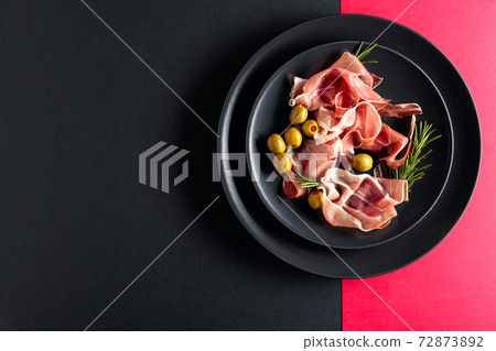 Prosciutto with rosemary and green olives on a black plate. Top view. 72873892