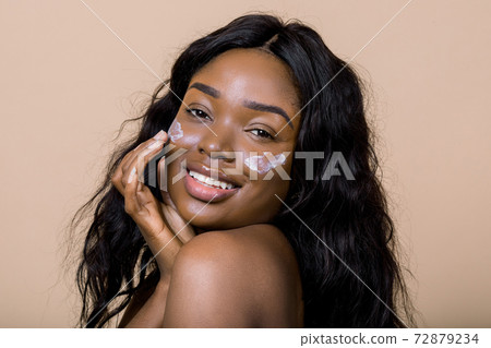 Beautiful naked black african model with flawless skin and long hair, applying moisturiser face cream on her cheeks, posing to camera with smile against beige background. Beauty, cosmetics, skincare Beautiful naked black african model with flawless skin and long hair, applying moisturiser face cream on her cheeks, posing to camera with smile against beige background. Beauty, cosmetics, skincare 72879234