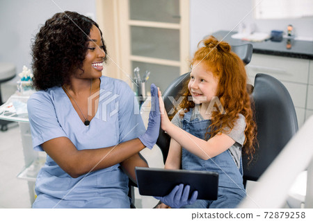 Medicine, pediatric dentistry and oral care concept. Happy female African dentist and curly red haired kid girl, having fun and giving high five, while holding tablet pc computer 72879258