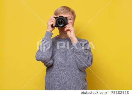 Mature female photographer with photocamera by face standing on yellow background. 72882528
