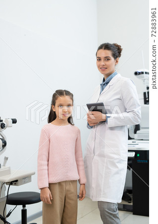 Happy young female ophthalmologist in whitecoat with tablet and schoolgirl 72883961