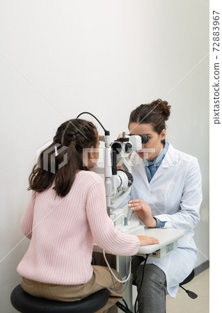 Young ophthalmologist in whitecoat checking eyesight of elementary schoolgirl Young ophthalmologist in whitecoat checking eyesight of elementary schoolgirl 72883967