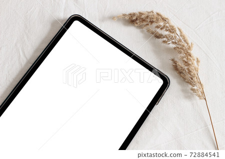 Closeup of tablet with dry grass on table. Blank, empty screen. White linen table cloth background in sunlight. Electronic device mock-up. Modern still life. Flat lay, top view. 72884541