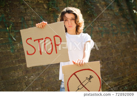 Young protesting woman in white shirt and jeans holds protest sign broadsheet placard with slogan 'Stop' and syringe sing as a symbol of vaccine for public demonstration on wall background Young protesting woman in white shirt and jeans holds protest sign broadsheet placard with slogan 'Stop' and syringe sing as a symbol of vaccine for public demonstration on wall background 72892009