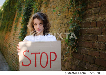 Young protesting woman in white shirt and jeans holds protest sign broadsheet placard with slogan 'Stop' for public demonstration on wall background. Young protesting woman in white shirt and jeans holds protest sign broadsheet placard with slogan 'Stop' for public demonstration on wall background. 72892010