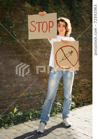 Young protesting woman in white shirt and jeans holds protest sign broadsheet placard with slogan Stop and syringe sing as a symbol of vaccine for public demonstration on wall background Young protesting woman in white shirt and jeans holds protest sign broadsheet placard with slogan Stop and syringe sing as a symbol of vaccine for public demonstration on wall background 72892063