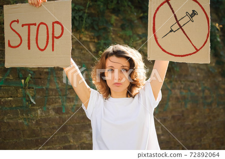 Young protesting woman in white shirt and jeans holds protest sign broadsheet placard with slogan 'Stop' and syringe sing as a symbol of vaccine for public demonstration on wall background 72892064