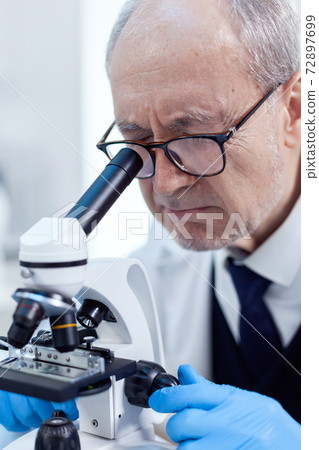 Elderly aged scientist in modern facility looking through a microscope Elderly aged scientist in modern facility looking through a microscope 72897699