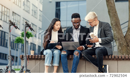 Elegant multiracial colleagues sitting on the bench near office and discussing business matters outdoors. Elegant multiracial colleagues sitting on the bench near office and discussing business matters outdoors. 72902504