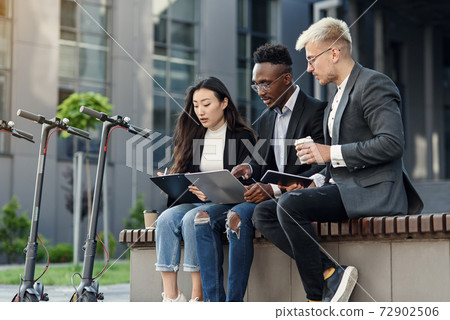 Elegant multiracial colleagues sitting on the bench near office and discussing business matters outdoors. Elegant multiracial colleagues sitting on the bench near office and discussing business matters outdoors. 72902506