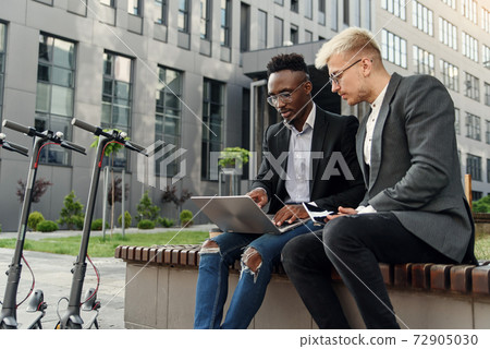 Elegant multiracial colleagues sitting on the bench near office and discussing business matters outdoors. Elegant multiracial colleagues sitting on the bench near office and discussing business matters outdoors. 72905030