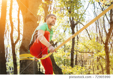 A bearded man in age balances while sitting on a taut slackline in the autumn forest. Outdoor Leisure 72905990