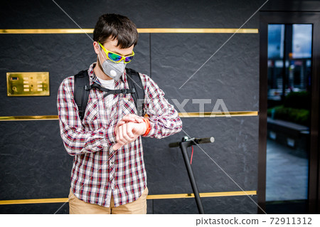 Courier uses electric scooter in city. Young attractive Caucasian man with backpack, stylishly dressed, using mask against smog and air pollution in city, waiting customer and looking at wristwatch Courier uses electric scooter in city. Young attractive Caucasian man with backpack, stylishly dressed, using mask against smog and air pollution in city, waiting customer and looking at wristwatch 72911312