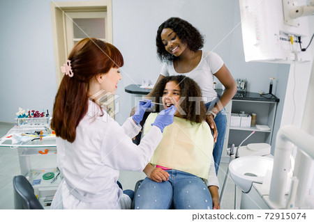 Young smiling African woman mother with her cute preteen daughter, visit their pediatric dentist for regular teeth checkup at stomatology clinic. Child with mom at dentist's office Young smiling African woman mother with her cute preteen daughter, visit their pediatric dentist for regular teeth checkup at stomatology clinic. Child with mom at dentist's office 72915074