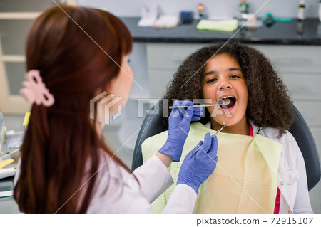 Close up view of pretty kid, african american curly girl in dentist chair, with mouth open, having dental examination. Back view of female dentist in gloves, holding tools 72915107