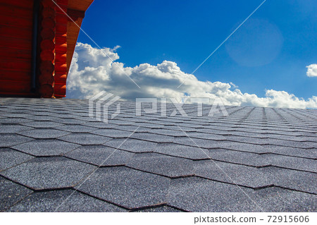 Gray soft roof on the roof of the house. Cloudy blue sky on background. Gray soft roof on the roof of the house. Cloudy blue sky on background. 72915606