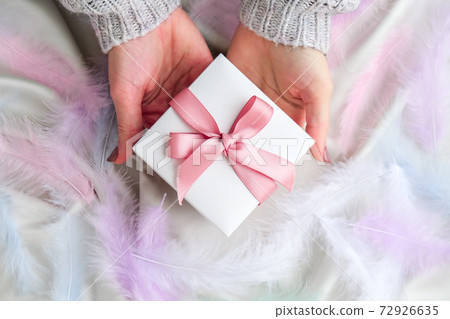 Female hand holding White gift box with pink ribbon on white silk texture background. Colorful bird feather. Holiday concept. Female hand holding White gift box with pink ribbon on white silk texture background. Colorful bird feather. Holiday concept. 72926635