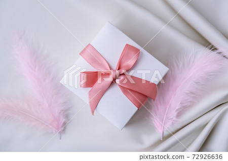 White gift box with pink ribbon on white silk texture background. Colorful bird feather. Holiday concept. White gift box with pink ribbon on white silk texture background. Colorful bird feather. Holiday concept. 72926636