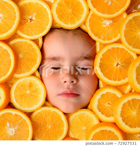 The face of a beautiful little girl is surrounded by pieces of sliced orange, orange useful fruit rich in vitamin C. 72927531