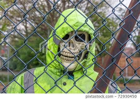 Skull of skeleton in green high-visibility jacket and hood, seen through metal fence. 72930450
