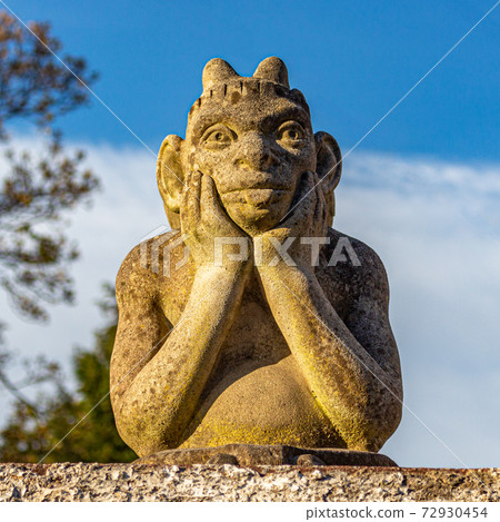 Stone gargoyle or goblin with head in hands, looking over garden wall, with blue sky in background. Stockport, Cheshire, UK. Stone gargoyle or goblin with head in hands, looking over garden wall, with blue sky in background. Stockport, Cheshire, UK. 72930454