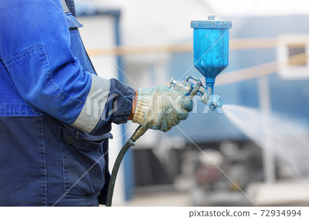 Spray gun in hand. A worker in overalls and a glove holds a spray compressor for painting in his 72934994