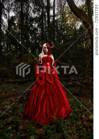 Maleficent Woman in Red Clothing and Horns in dark Forest. Posing in magik forest Maleficent Woman in Red Clothing and Horns in dark Forest. Posing in magik forest 72937783