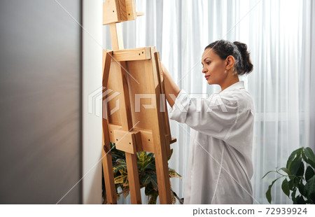 Young beautiful female artist wearing white shirt stands in front of easel and looks down 72939924