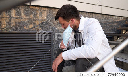 Serious doctor resting on stairs after very long shift in hospital. Tired doctor in mask, white uniform and stethoscope resting while sit on staircase outside clinic, stress headache and rubbing eyes 72940900