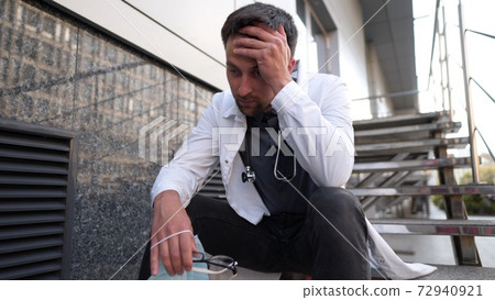 Worried and tired doctor sitting on stairs outside hospital. Exhausted physician in white lab coat and medical protective mask, sit on staircase clinic. Stress healthcare worker during COVID-19 72940921