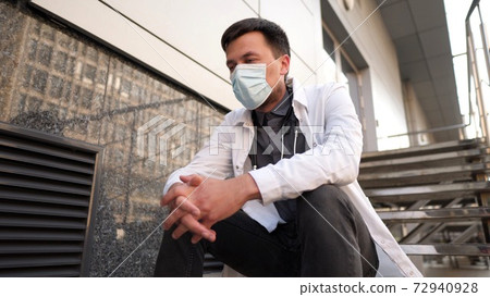 Serious doctor resting on stairs after very long shift in hospital. Tired doctor in mask, white uniform and stethoscope resting while sit on staircase outside clinic, stress headache and rubbing eyes Serious doctor resting on stairs after very long shift in hospital. Tired doctor in mask, white uniform and stethoscope resting while sit on staircase outside clinic, stress headache and rubbing eyes 72940928