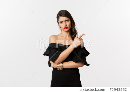 Fashion and beauty. Skeptical glamour woman with red lips, black dress, pointing finger right at something lame and boring, standing over white background 72943262