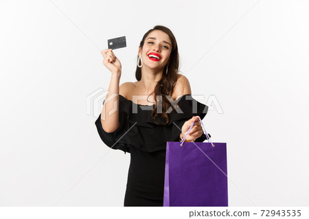 Stylish young woman in black dress going shopping, holding bag and credit card, smiling pleased, standing over white background Stylish young woman in black dress going shopping, holding bag and credit card, smiling pleased, standing over white background 72943535