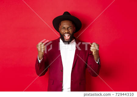 Satisfied lucky Black man celebrating victory, screaming from rejoice, winning prize, saying yes and fist pump, red background Satisfied lucky Black man celebrating victory, screaming from rejoice, winning prize, saying yes and fist pump, red background 72944310