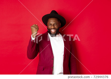 Fashion and party concept. Excited Black man rejoicing of winning, making fist pump gesture and say yes with satisfaction, standing against red background 72944376