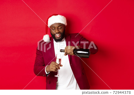 Christmas, party and holidays concept. Handsome Black man in santa hat pouring glass of champagne and smiling, celebrating New Year, red background Christmas, party and holidays concept. Handsome Black man in santa hat pouring glass of champagne and smiling, celebrating New Year, red background 72944378