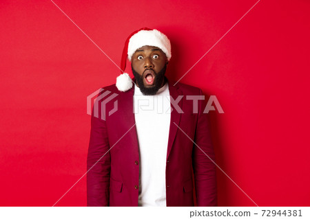 Christmas, party and holidays concept. Shocked Black man in santa hat drop jaw and stare at camera, stand against red background Christmas, party and holidays concept. Shocked Black man in santa hat drop jaw and stare at camera, stand against red background 72944381