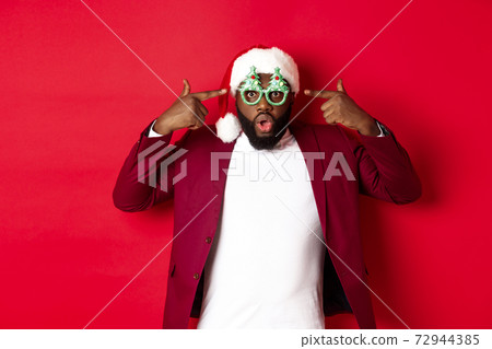 Merry Christmas. Cheerful Black man wearing funny party glasses and santa hat, smiling joyful, celebrating winter holidays, standing over red background Merry Christmas. Cheerful Black man wearing funny party glasses and santa hat, smiling joyful, celebrating winter holidays, standing over red background 72944385