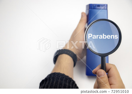 Chemical components on the shampoo label: parabens . A hand holds a blue jar and a magnifier, where the harmful ingredients of a detergent are written in close up. 72951010