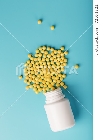 Yellow vitamin C capsules spilled out of a white jar against a blue background. 72951521