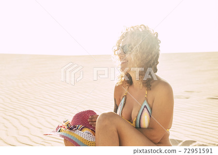 Sunny day of vacation at the beach -  cheerful happy beautiful middle age young caucasian woman sit down on the sand with coloured bikini enjoying the sun and the summer holiday lifestyle 72951591