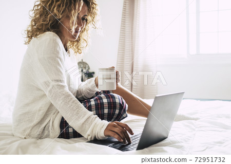 Young adult woman working at home in the bedroom during morning breakfast work activity - quarantine for coronavirus health emergency - happy female with computer laptop and coffe cup - bright white 72951732