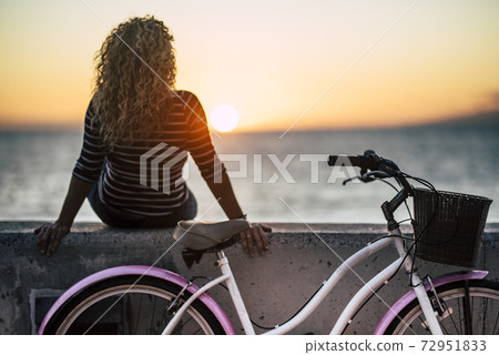 People enjoying outdoor leisure actvity looking a beautiful coloured sunset - woman with long curly blonde hair viewed from back enjoy the relax after a bike tour - horizon and sun with ocean People enjoying outdoor leisure actvity looking a beautiful coloured sunset - woman with long curly blonde hair viewed from back enjoy the relax after a bike tour - horizon and sun with ocean 72951833