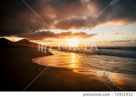 Wonderful timeless sunset at the beach with dramatic clouds and waves - mountains in background and wild scenic place - golden and fire colors in a beauty of nature 72952932