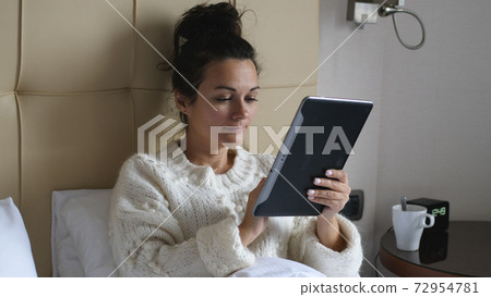 Young pretty woman freelancer is sitting on a bed drinking coffee and working on a screen tablet, morning time, remote work from home. 72954781